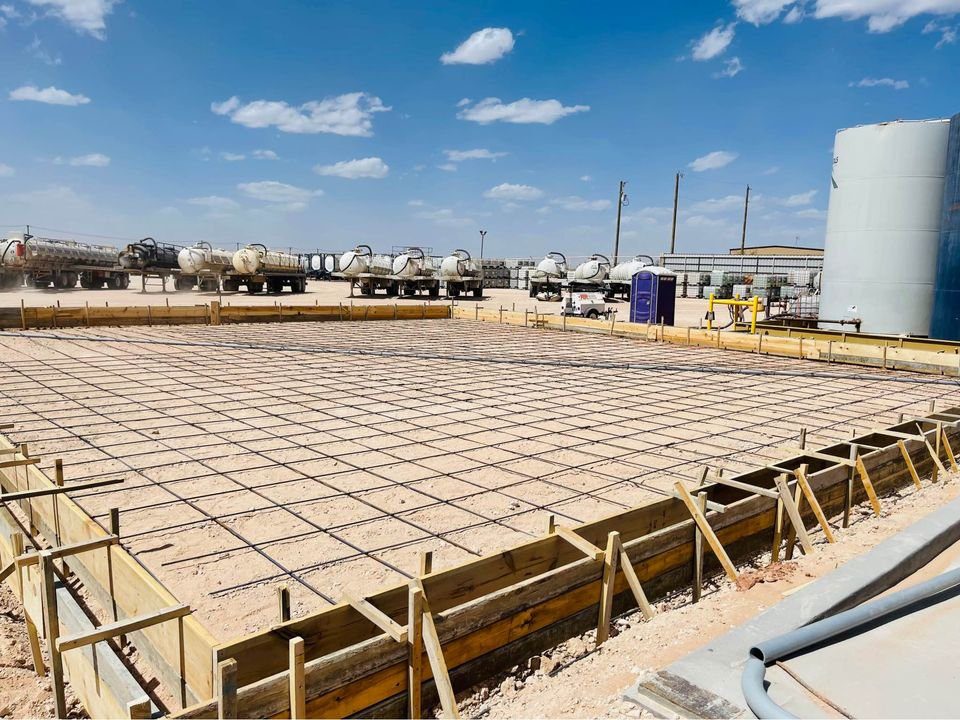 A construction site in Mesa, AZ features a large, rectangular foundation being prepped by commercial concrete contractors. Wooden boards frame the area, lined with a grid of metal rebar. Trucks and a portable toilet dot the background under a blue sky with scattered clouds.