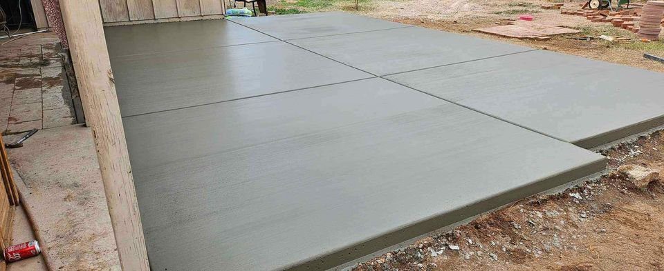 A freshly poured concrete slab with a smooth finish, divided into six rectangular sections near a building in Mesa, AZ. The surrounding area reveals dirt and construction materials, evidence of the skilled craftsmanship of the foundation contractor at work.