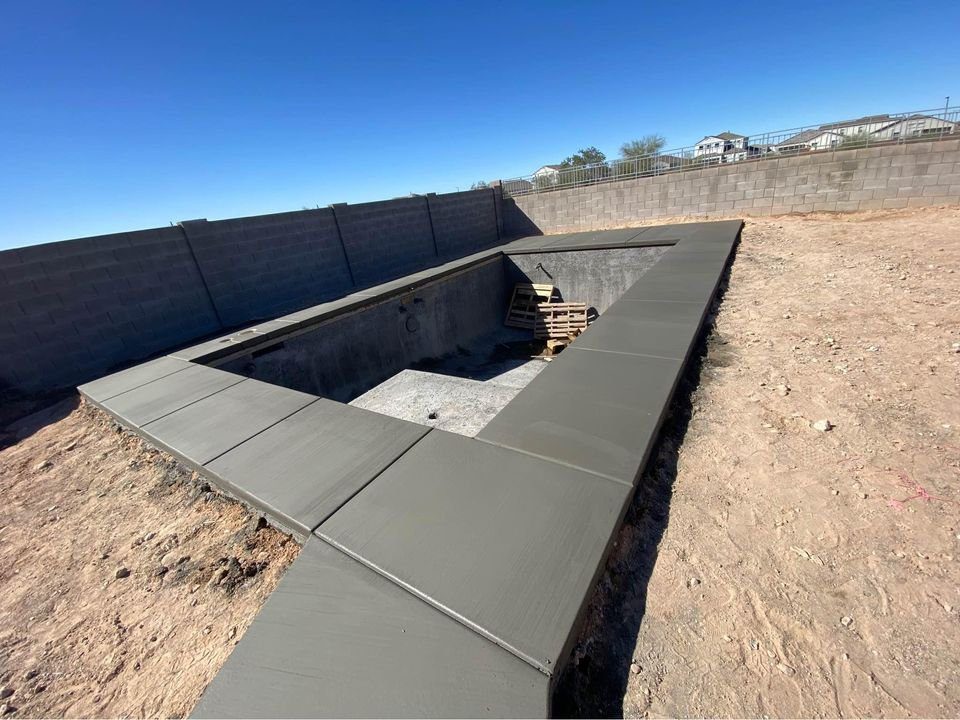 An empty swimming pool under construction, surrounded by a concrete pool deck in Mesa, AZ. A pallet rests at the deep end, with the area bordered by a tall brick wall under a clear blue sky.
