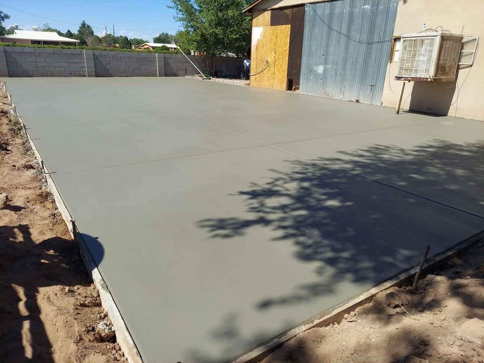 A concrete slab, freshly poured by a foundation contractor in Mesa, AZ, lies in a backyard, bordered by wooden forms. Adjacent to the building with its corrugated metal wall and air conditioning unit, the slab is dappled with the shadows of nearby trees.