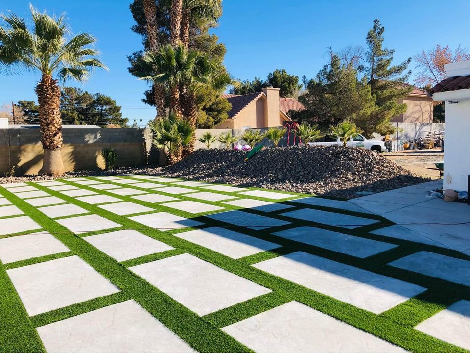 A backyard with a geometric design featuring alternating squares of artificial grass and concrete, skillfully laid out by Mesa Master Concrete. Palm trees and a large pile of rocks are visible in the background, with a clear blue sky overhead.