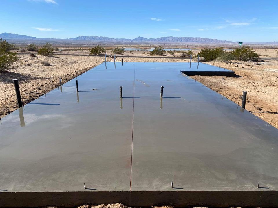 A freshly poured Mesa Master Concrete slab is curing under a clear blue sky in a desert landscape. Surrounding the slab are patches of dry soil and sparse desert vegetation. Mountains are visible in the distance, highlighting the expertise of premier concrete contractors in such rugged terrains.