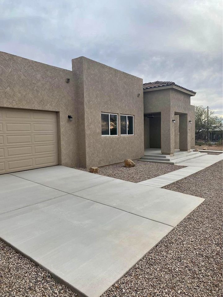 A modern, single-story house with a flat roof and stucco exterior features a beige garage door and a concrete driveway crafted by expert concrete contractors. The front entrance is accented with a small porch, and the surrounding landscape is artistically covered in gravel.