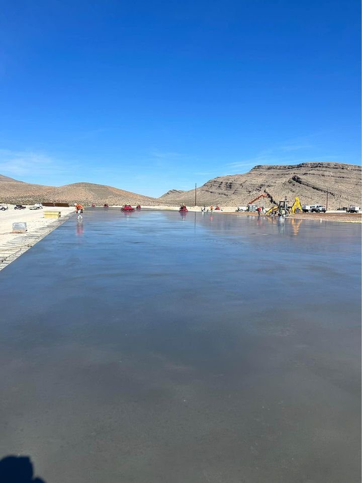 A smooth concrete slab stretches out in a barren landscape under the clear blue skies of Mesa, AZ. Workers and construction equipment, including cranes and vehicles from commercial concrete contractors, are visible in the distance with mountains in the background.