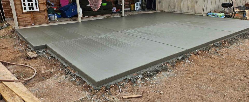 Freshly poured concrete slab with a smooth surface and defined edges sits amidst bare earth and construction materials in Mesa, AZ. A wooden structure is partially visible in the background, hinting at the meticulous work of a skilled foundation contractor.