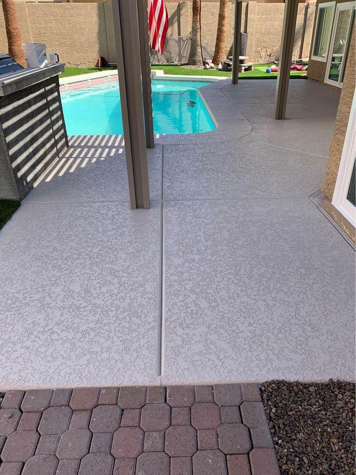 A backyard in Mesa, AZ, features a swimming pool surrounded by a smooth concrete pool deck. An American flag is hung under a pergola. Pavers and stones lead to the pool area, creating a sunny and relaxed setting ideal for enjoying warm days.