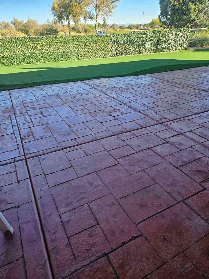A wet, red-tiled patio extends towards a well-manicured green lawn, framed by a lush hedge and tall trees under a clear blue sky. Crafted with precision by Mesa Master Concrete, a hint of a white chair is visible on the left side of the image.