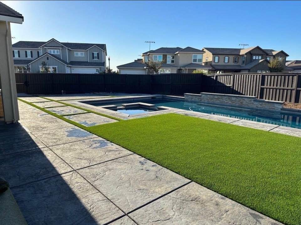 A backyard in Mesa, AZ features a rectangular swimming pool and hot tub on a sleek concrete patio. Artificial grass lies between the pool and house, while the backdrop of suburban homes peeks over a dark wooden fence, showcasing the work of top-notch concrete contractors.