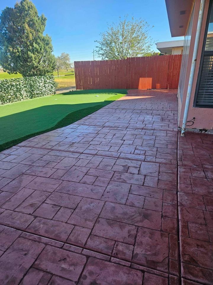 A concrete patio featuring a decorative pattern sits beside a synthetic grass area with a putting green. Surrounded by a wooden fence and trees, this project showcases the expertise of licensed concrete contractors. Contact us for your free quote today!.